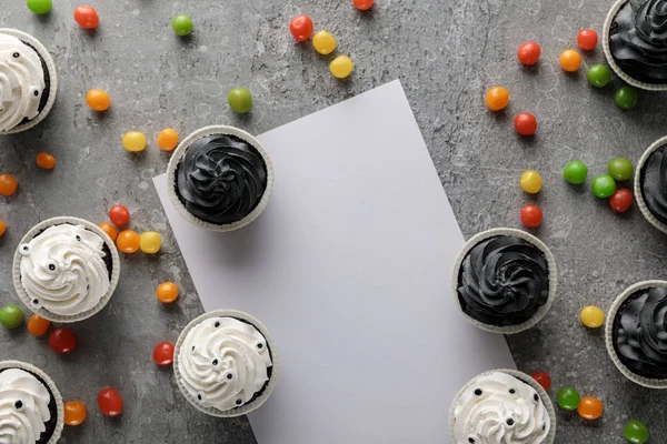 Vista dall'alto di deliziosi cupcake di Halloween con bonbon colorati sparsi vicino alla carta bianca su una superficie grigia in cemento — Foto stock