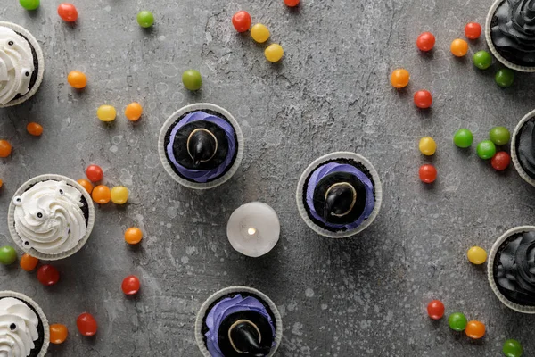 Draufsicht auf köstliche Halloween-Cupcakes und brennende Kerze mit verstreuten bunten Bonbons auf betongrauer Oberfläche — Stockfoto