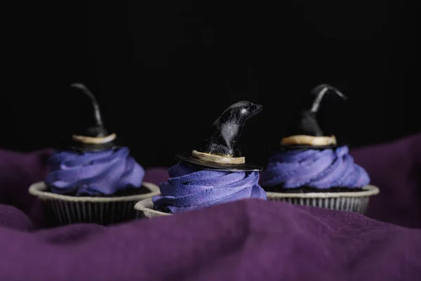 Saborosos cupcakes de Halloween com creme azul e chapéus de bruxa decorativos em pano roxo isolado em preto — Fotografia de Stock