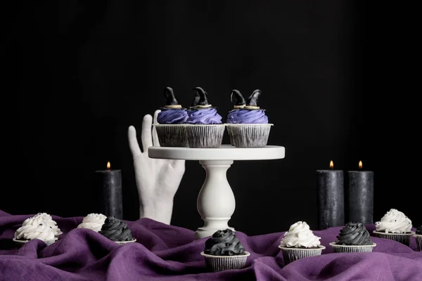 Tasty Halloween cupcakes on white stand near burning candles and decorative hand on purple cloth isolated on black — Stock Photo