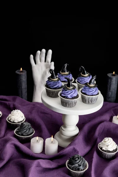 Leckere Halloween-Cupcakes auf weißem Ständer neben brennenden Kerzen und dekorative Hand auf lila Tuch isoliert auf schwarz — Stockfoto