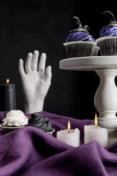 Tasty Halloween cupcakes on white stand near burning candles and decorative hand on purple cloth isolated on black — Stock Photo