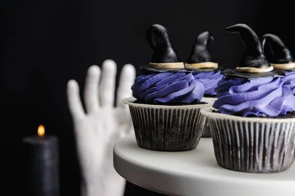 Foco selectivo de sabrosos cupcakes de Halloween con crema azul y sombreros de bruja decorativos en soporte blanco aislado en negro - foto de stock