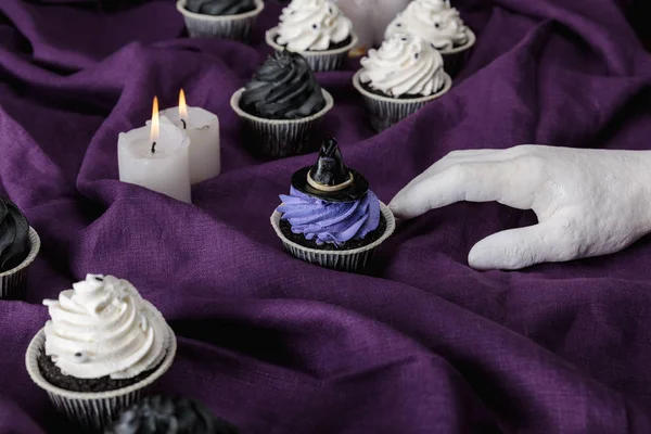 Dekorative Hand in der Nähe von leckeren Halloween-Cupcakes und brennenden Kerzen auf violettem Tuch — Stockfoto