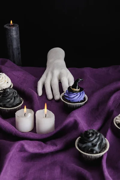 Dekorative Hand in der Nähe von leckeren Halloween-Cupcakes und brennenden Kerzen auf lila Tuch isoliert auf schwarz — Stockfoto