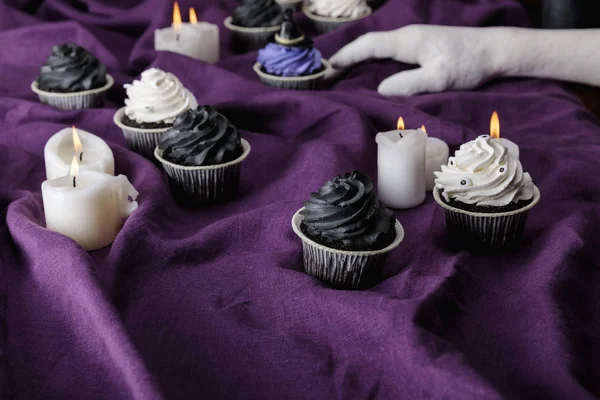 Dekorative Hand in der Nähe von leckeren Halloween-Cupcakes in der Nähe brennender Kerzen auf lila Tuch — Stockfoto