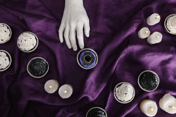 Top view of decorative hand near tasty Halloween cupcake with blue cream and witch hat near burning candles on purple cloth — Stock Photo