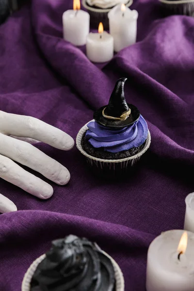 Mão decorativa perto de saboroso cupcake Halloween com creme azul e chapéu de bruxa decorativo perto de velas ardentes em pano roxo — Fotografia de Stock