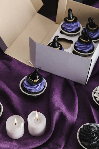 Savoureux cupcakes d'Halloween avec crème bleue et chapeaux de sorcière décoratifs en boîte près de bougies brûlantes sur tissu violet — Photo de stock