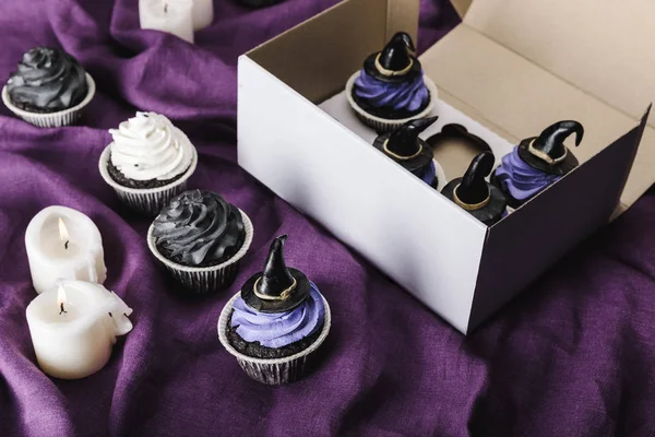 Вкусные кексы на Хэллоуин с голубым кремом и декоративные колпаки в коробке возле горящих свечей на фиолетовой ткани — стоковое фото