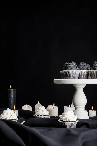 Leckere Halloween-Cupcakes mit schwarzer Sahne auf weißem Ständer in der Nähe brennender Kerzen isoliert auf schwarz — Stockfoto