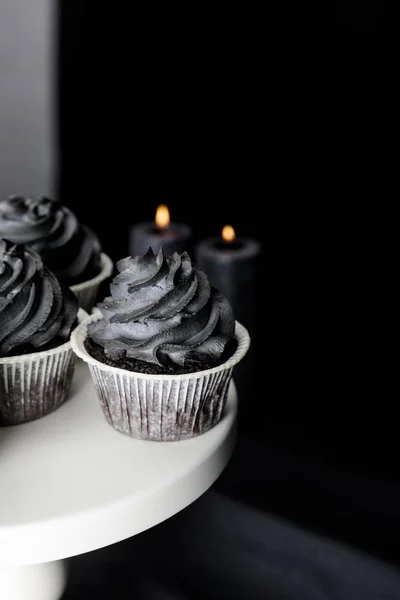 Fuoco selettivo di gustosi cupcake di Halloween con crema nera sullo stand vicino a candele accese isolate sul nero — Foto stock