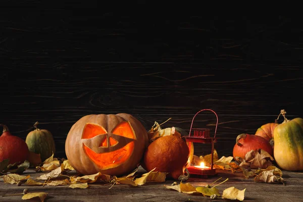 Calabaza de Halloween espeluznante tallada, hojas otoñales y vela ardiente sobre mesa rústica de madera sobre fondo negro — Stock Photo