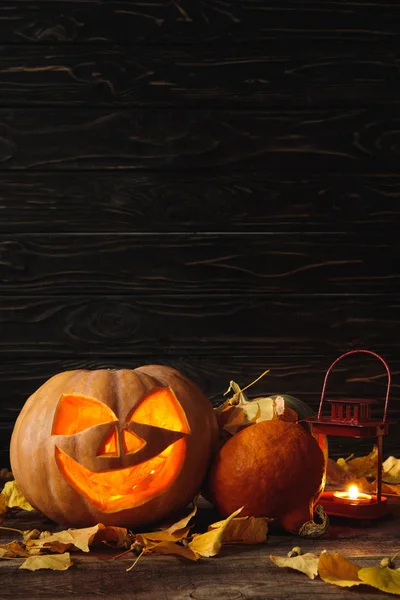 Zucca di Halloween spettrale intagliato, foglie autunnali e candela ardente su tavolo rustico in legno su sfondo nero — Foto stock