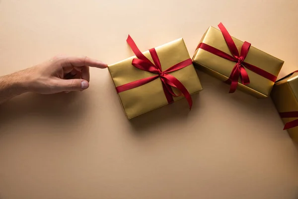 Vue recadrée de l'homme touchant des boîtes-cadeaux dorées avec des rubans rouges sur fond beige — Photo de stock