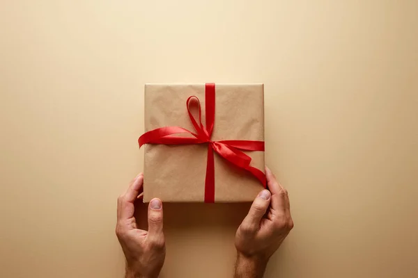 Обрізаний вид людини, що тримає подарункову коробку в ремісничому папері з червоною стрічкою на бежевому фоні — стокове фото