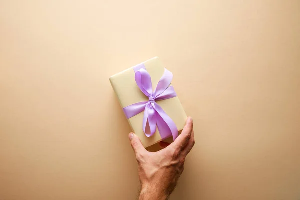 Cropped view of man holding gift box with violet ribbon on beige background — Stock Photo