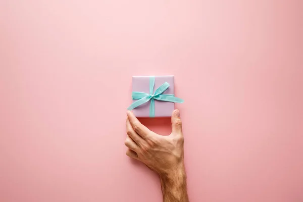 Cropped view of man holding violet small gift box with blue satin ribbon on pink background — Stock Photo