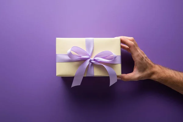 Cropped view of man holding gift box with violet ribbon on purple background — Stock Photo