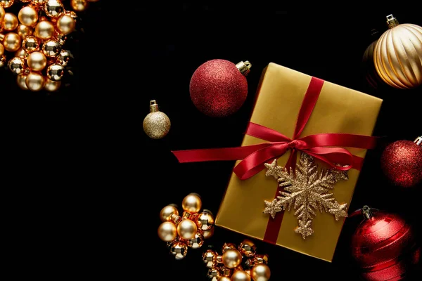 Vista dall'alto del regalo di Natale dorato lucido con nastro rosso e fiocco di neve vicino alle bagattelle isolate sul nero — Foto stock