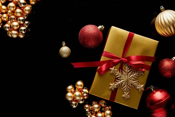 Top view of shiny golden Christmas gift with red ribbon and snowflake near baubles isolated on black — Stock Photo