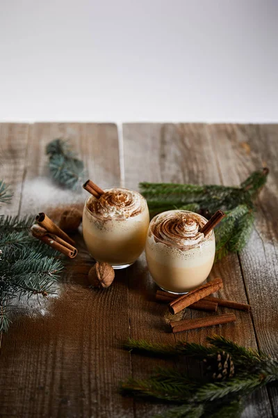 Eggnog cocktail with whipped cream and cinnamon near spruce branches and powdered sugar scattered on wooden table isolated on grey — Stock Photo