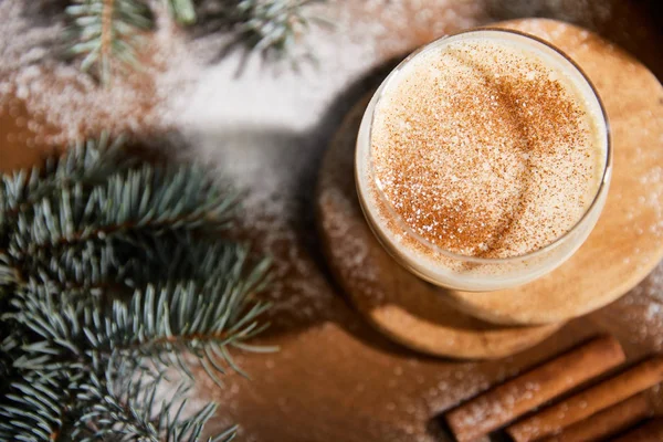 Blick von oben auf köstlichen Eierlikör-Cocktail auf rundem Holzbrett in der Nähe von Fichtenzweigen und verstreutem Zuckerpulver — Stockfoto