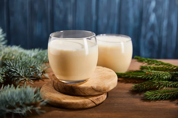 Delicious eggnog cocktail on round wooden boards near spruce branches on blue textured background — Stock Photo