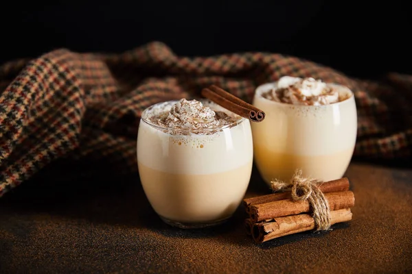 Delicioso cóctel de ponche de huevo, ramas de canela y paño a cuadros en la mesa cubierta con polvo de canela aislado en negro - foto de stock