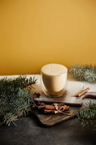 Traditional eggnog cocktail on chopping board covered with sugar powder, spruce branches and cinnamon sticks on orange background — Stock Photo