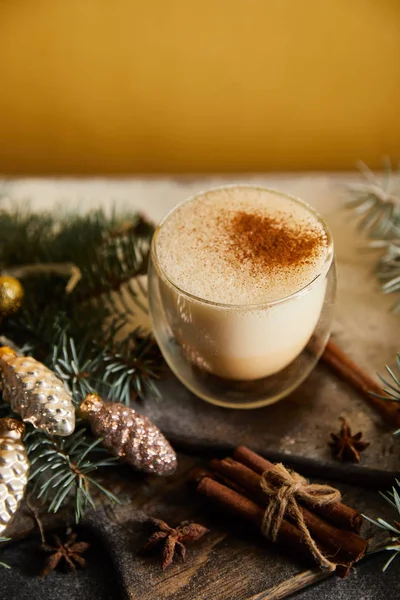 Eggnog cocktail on chopping board covered with sugar powder, spruce branches, cinnamon sticks and Christmas baubles on orange background — Stock Photo