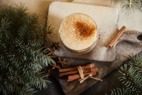 Draufsicht auf aromatisierten Eierlikör-Cocktail auf Schneidebrett mit Zuckerpuder, Zimtstangen und Fichtenzweigen — Stockfoto