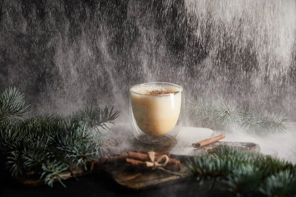 Tasty eggnog cocktail on cutting board near spruce branches and cinnamon sticks on black background with powdered sugar falling like snow — Stock Photo