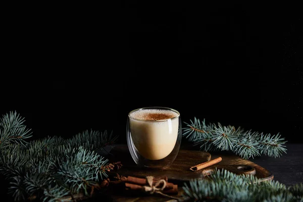 Delicious eggnog cocktail on cutting board near spruce branches and cinnamon sticks isolated on black — Stock Photo