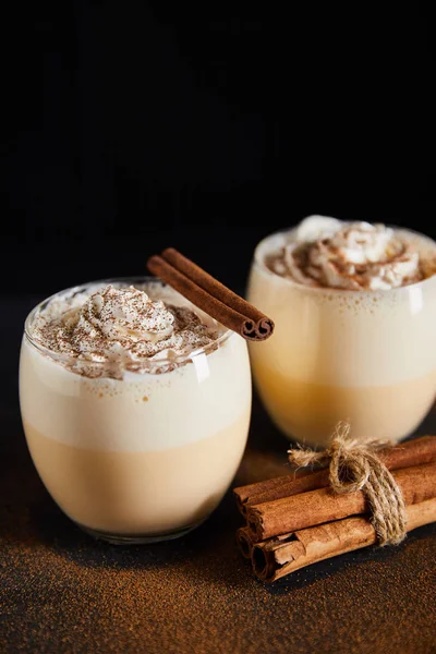 Délicieux cocktail au lait de poule avec crème fouettée et bâtonnets de cannelle sur la table recouvert de poudre de cannelle isolé sur noir — Photo de stock