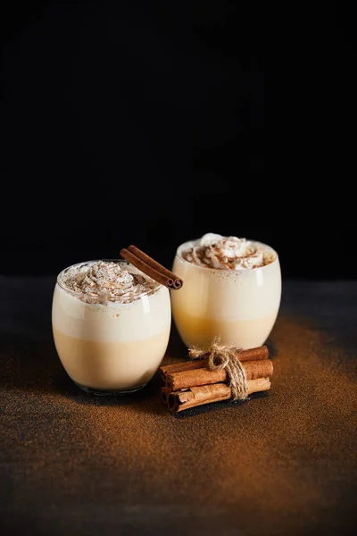 Delicioso cóctel de ponche de huevo con crema batida y palitos de canela en la mesa cubierta con polvo de canela aislado en negro - foto de stock