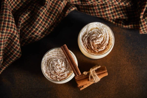 Top view of eggnog cocktail with whipped cream, cinnamon sticks and checkered cloth on table covered with cinnamon powder — Stock Photo