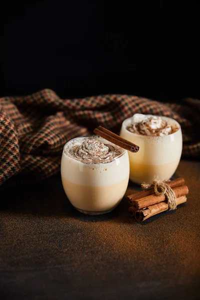 Eggnog cocktail with whipped cream, cinnamon sticks and checkered cloth on table covered with cinnamon powder isolated on black — Stock Photo