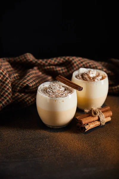 Cóctel de ponche de huevo con crema batida, palitos de canela y paño a cuadros en la mesa cubierta con polvo de canela aislado en negro - foto de stock