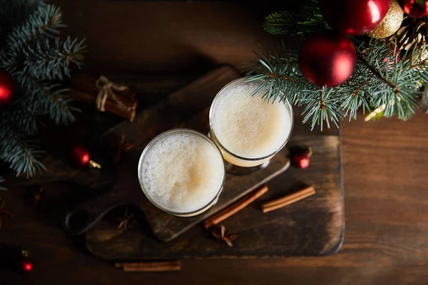 Draufsicht auf Eierlikör-Cocktail, Schneidebrett, Zimtstangen, Fichtenzweige und Weihnachtskugeln auf Holztisch — Stockfoto