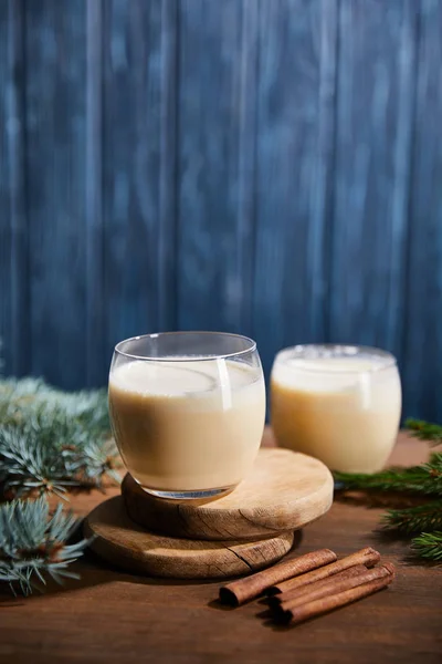 Leckerer Eierlikör-Cocktail auf runden Holzbrettern in der Nähe von Fichtenzweigen und Zimtstangen auf blau strukturiertem Hintergrund — Stockfoto