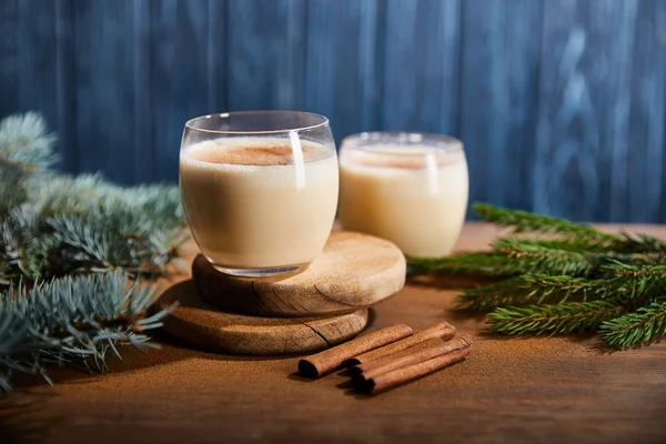 Tasty eggnog cocktail on round wooden boards near spruce branches and cinnamon sticks on blue textured background — Stock Photo