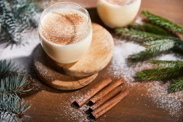 Flavored eggnog cocktail on round boards near spruce branches and cinnamon sticks on wooden table covered with sugar powder — Stock Photo