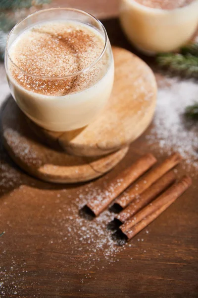 Cóctel de ponche de huevo con sabor en tablas redondas cerca de ramas de abeto y palitos de canela en la mesa de madera cubierta con azúcar en polvo - foto de stock