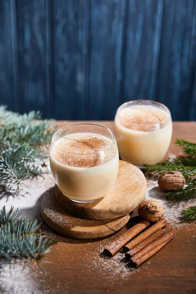 Delicioso cóctel de ponche de huevo, ramas de abeto, palitos de canela y nueces en la mesa de madera cubierta de azúcar en polvo sobre fondo de textura azul - foto de stock