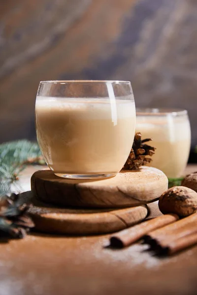 Enfoque selectivo de delicioso cóctel de ponche de huevo en tablas redondas, conos de abeto, palitos de canela y nueces sobre fondo de piedra de mármol - foto de stock