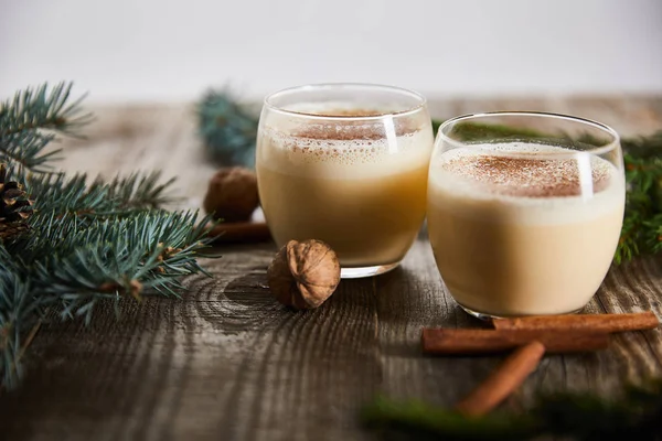 Focus selettivo di cocktail di zabaione aromatizzato vicino a bastoncini di cannella, noci e rami di abete rosso su tavolo in legno isolato su grigio — Foto stock