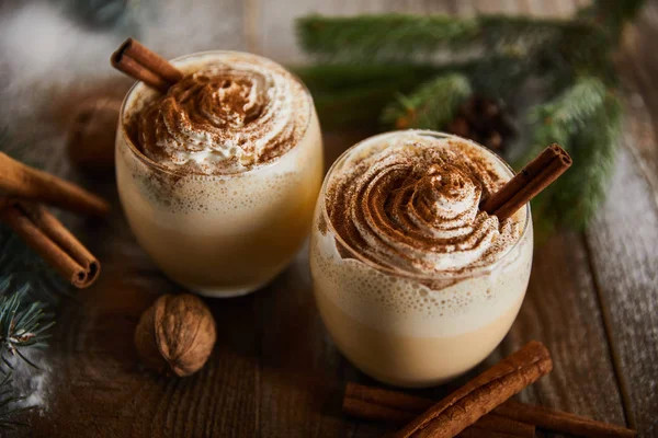 Traditional eggnog cocktail with whipped cream near cinnamon sticks, spruce branches and walnuts on wooden table — Stock Photo