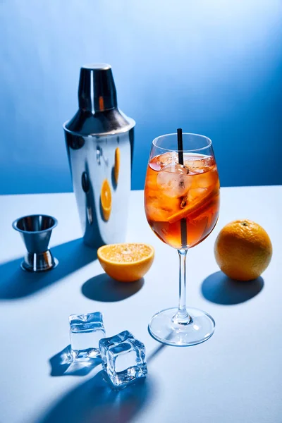 Cóctel Aperol Spritz, naranjas, coctelera, cubitos de hielo y taza de medir sobre fondo azul - foto de stock
