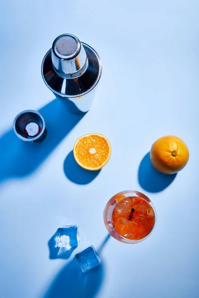 Вид сверху на коктейль Aperol Spritz, апельсины, шейкер, кубики льда и измерительная чашка на синем фоне — стоковое фото